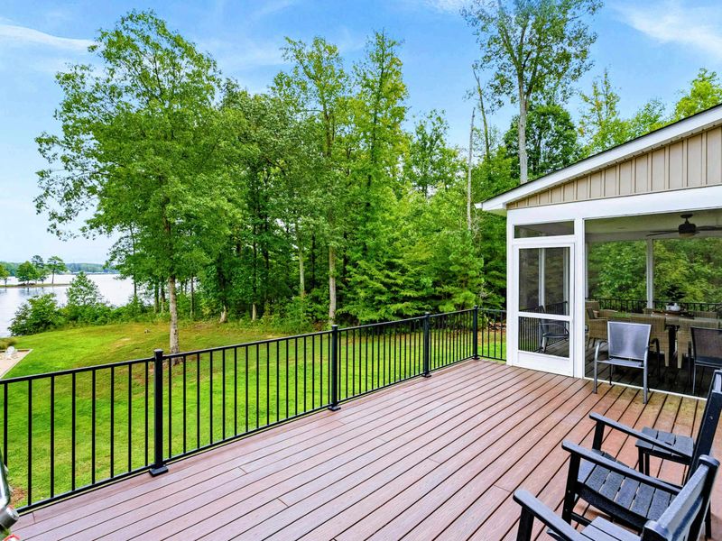 A spacious outdoor deck with a serene lake view, ample seating, a grill for entertaining, and access to a screened-in porch and lush green yard.