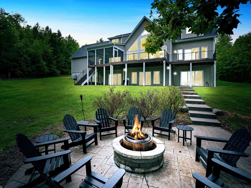 A spacious backyard with a fire pit seating area overlooking the lake, surrounded by greenery and showcasing a stunning multi-level lake house with large windows and outdoor spaces.