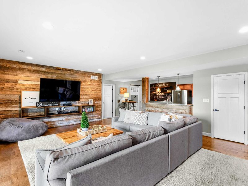 A spacious and inviting lower-level living area with a large sectional, rustic wood accent wall, entertainment center, and access to the backyard and lake views, complete with an adjacent bar and kitchenette.