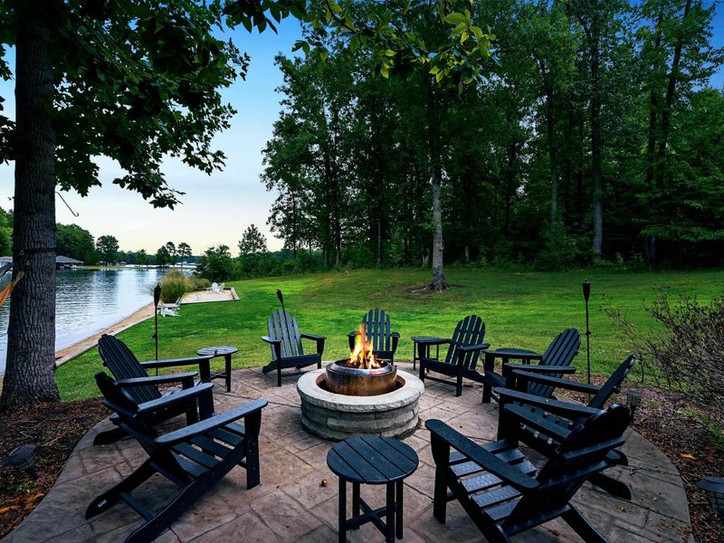 A spacious backyard with a fire pit seating area overlooking the lake, surrounded by greenery and showcasing a stunning multi-level lake house with large windows and outdoor spaces.