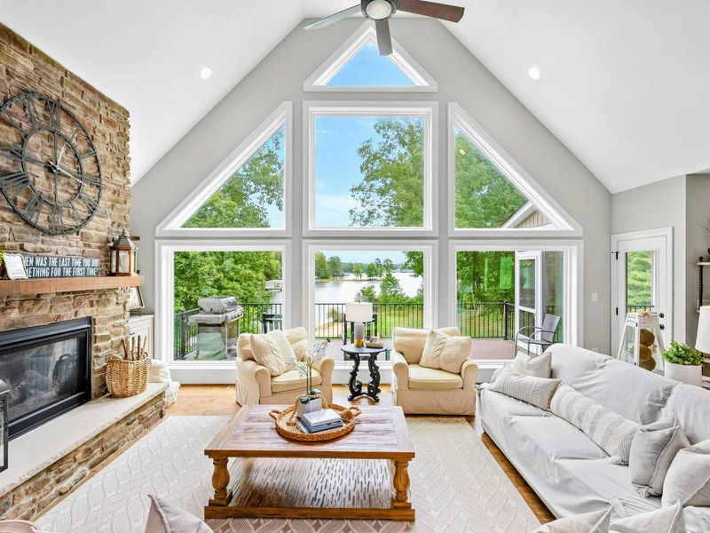 A bright and spacious lakefront living area with large floor-to-ceiling windows, cozy seating, a stone fireplace, and open access to the kitchen and outdoor deck.