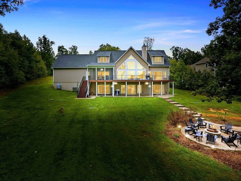 A spacious backyard with a fire pit seating area overlooking the lake, surrounded by greenery and showcasing a stunning multi-level lake house with large windows and outdoor spaces.