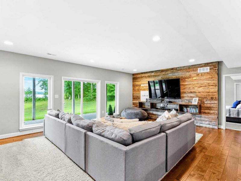 A spacious and inviting lower-level living area with a large sectional, rustic wood accent wall, entertainment center, and access to the backyard and lake views, complete with an adjacent bar and kitchenette.