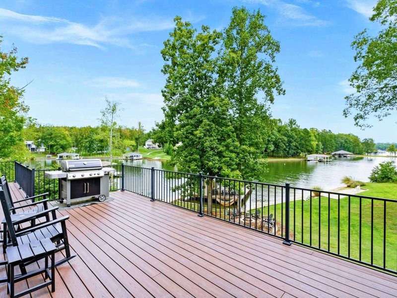 A spacious outdoor deck with a serene lake view, ample seating, a grill for entertaining, and access to a screened-in porch and lush green yard.