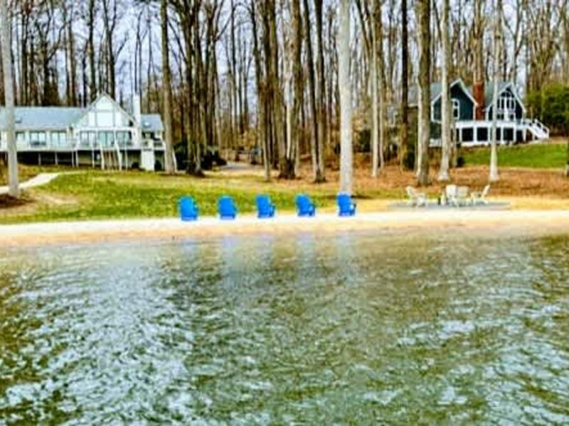 Private Sandy Shoreline: A peaceful view from the water reveals your private stretch of sandy beach, complete with colorful Adirondack chairs and a spacious, wooded backyard retreat on Lake Anna.