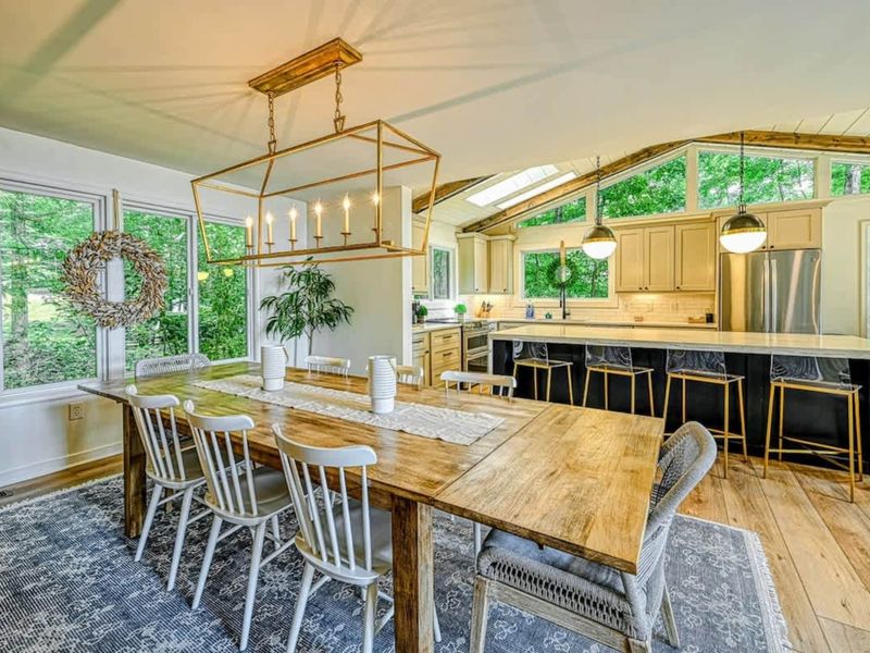 Dining and Kitchen: A rustic dining table with modern chairs sits under a gold chandelier, seamlessly connected to a sleek, light-filled kitchen with bar seating and large windows.