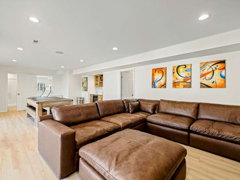Lower Level - Oversized brown leather sectional with colorful abstract triptych art, recessed lighting, and the game area behind.