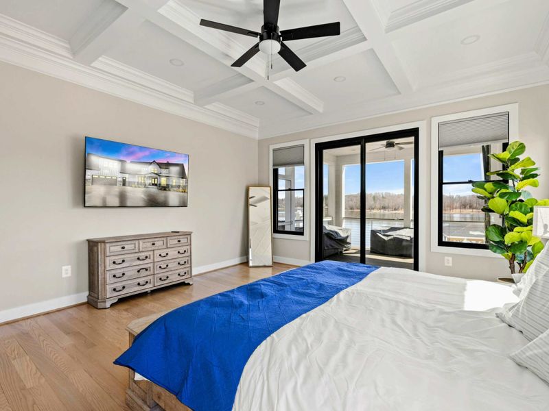 Primary Suite: Luxurious and spacious bedroom featuring a coffered ceiling, elegant decor, a mounted TV, and private access to a covered patio with stunning lake views.