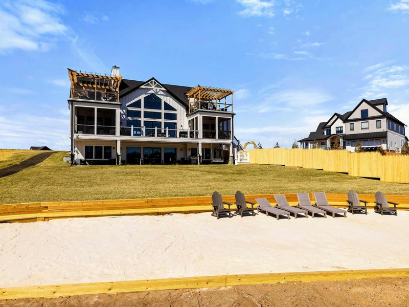 Luxury Lakefront Retreat: Stunning three-story lakefront home with expansive balconies, lush green lawn, and a private sandy beach featuring lounge chairs for ultimate relaxation by the water.