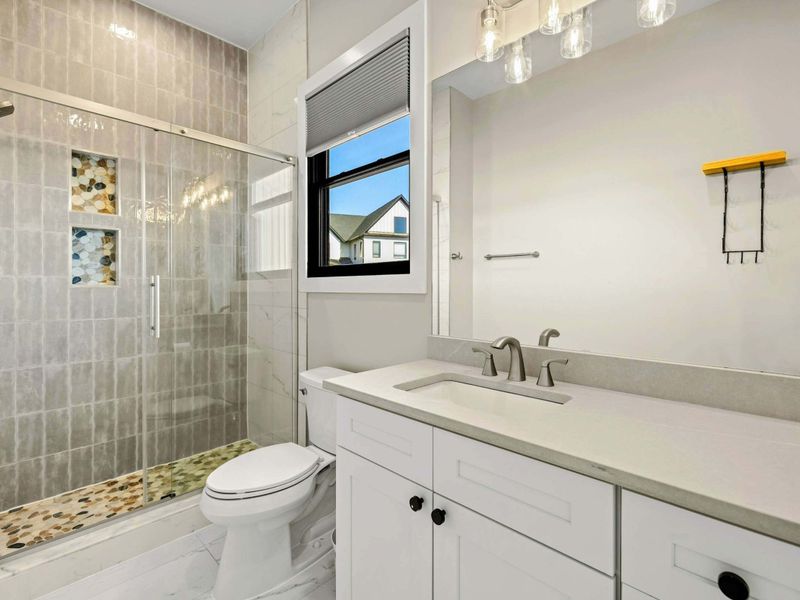 Suite #2 Bathroom: Elegant en-suite featuring a sleek quartz vanity, stylish lighting, and a spacious glass-enclosed shower with custom tile and pebble flooring for a spa-like experience.