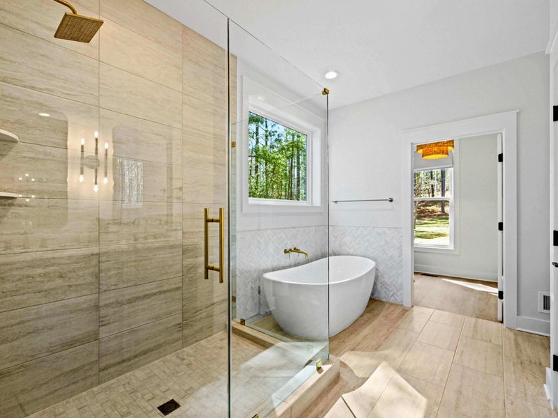 Spa Shower - A wood-tile walk-in shower with a gold rainhead anchors the primary bath, the freestanding soaking tub waiting just beyond.