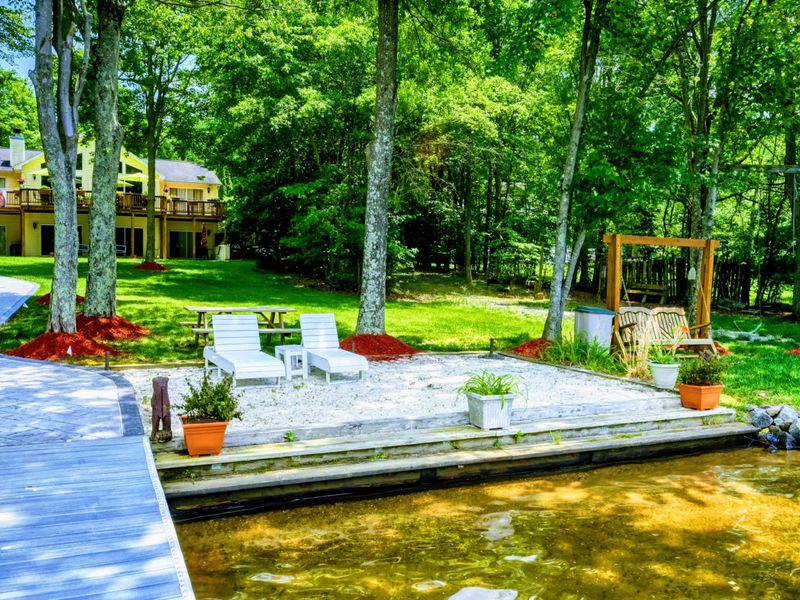 Sandy Lakeside Haven – Discover a serene spot right by the water, featuring a sandy stretch with comfortable seating, surrounded by towering trees. A charming place for leisure and lakeside chats at Lake Anna.