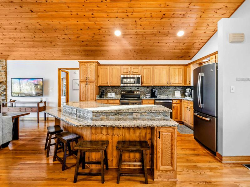 Modern Kitchen – This fully equipped kitchen features sleek granite countertops, a large center island with bar seating, and beautiful wooden cabinetry, all set under warm wooden ceilings. Ideal for preparing meals and casual dining.