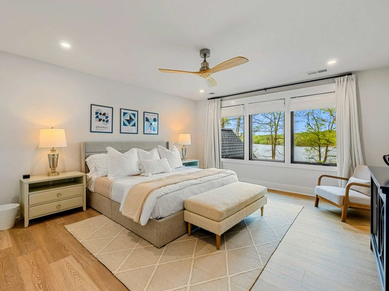 Primary Suite - King bed with plush white bedding, an upholstered headboard, and lake views through black-framed windows.