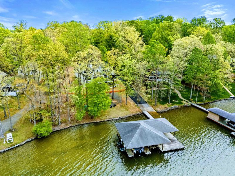 Aerial - The covered dock and waterfront lawn viewed from above, with white Adirondack chairs dotting the shoreline.