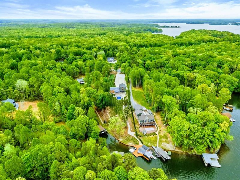 Aerial - Drone view of the waterfront neighborhood showing the home, dock, and tree-lined road leading to the water.