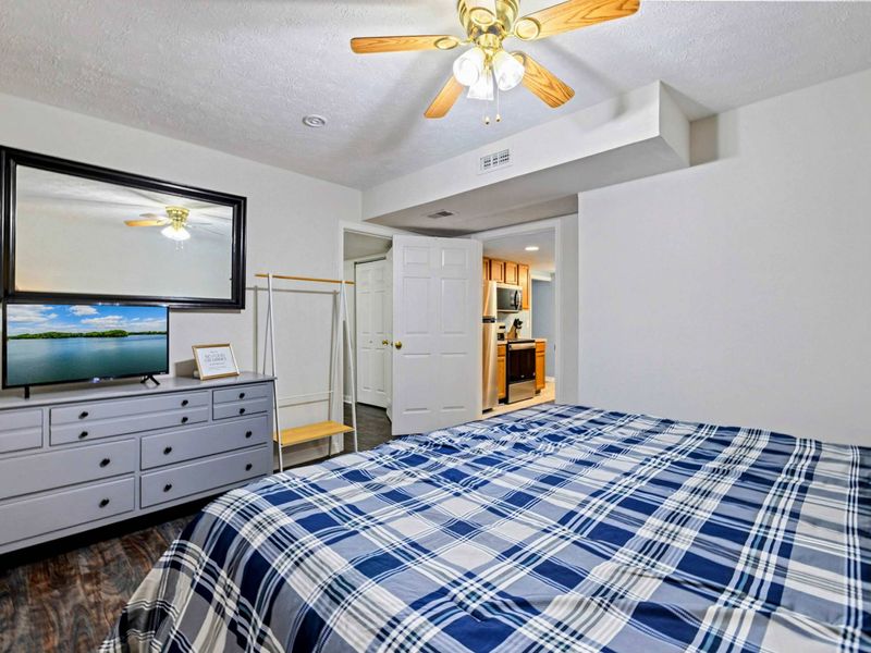 Bedroom - Alternate angle of the king bed showing the wall-mounted TV, gray dresser, clothing rack, and doorway to the kitchenette.