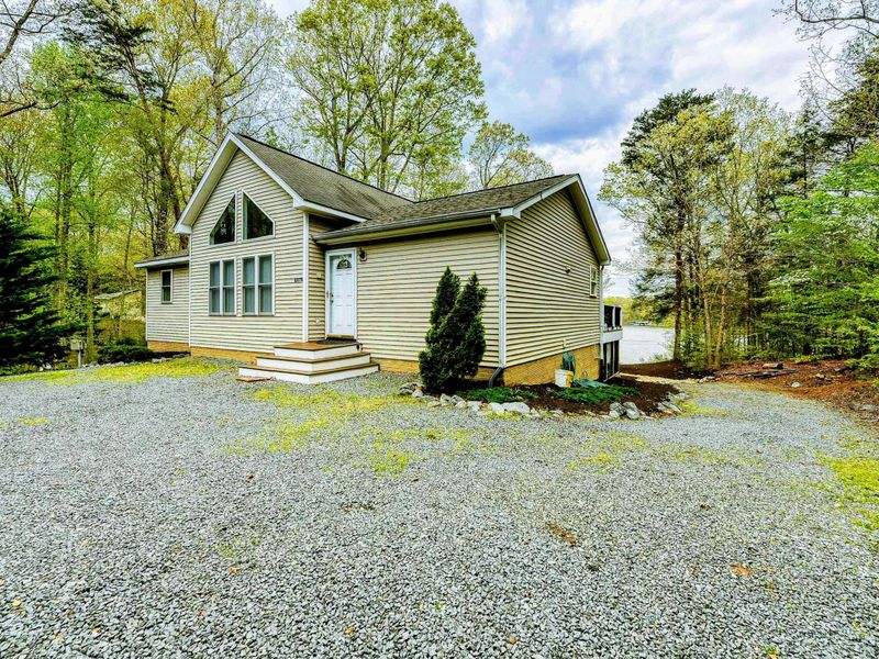 Exterior Front - Tan siding with A-frame peak, covered entry porch, and gravel parking among the tall pines.