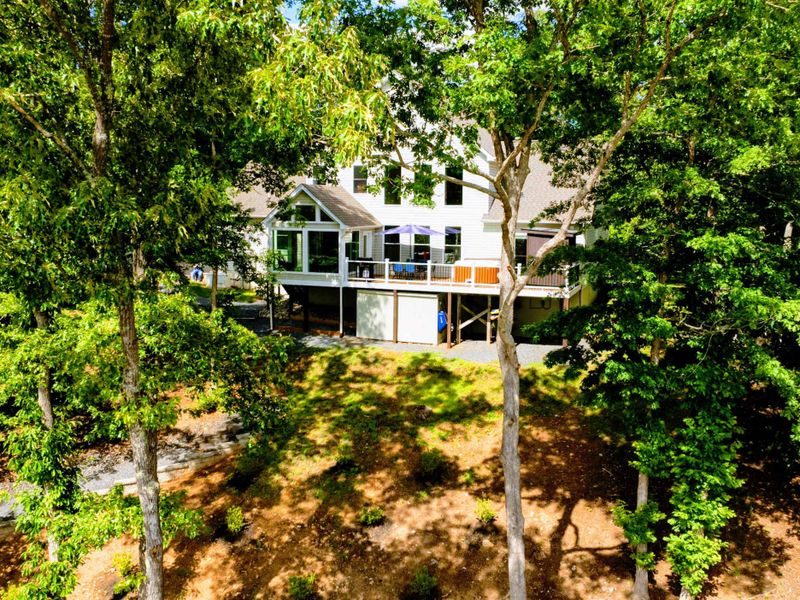 Aerial - Rear of the home nestled among mature trees with the deck, sunroom, and lakeside yard visible.