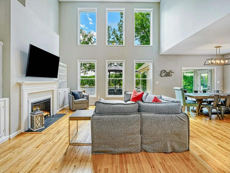 Living Room - Two-story great room with fireplace, wall-mounted TV, built-in shelving, gray sectional, and six tall windows.