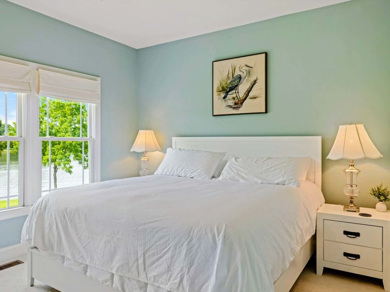 King Bedroom - A closer look at the white king bed, crystal lamps, and heron print, with the lake and green lawn framed through the window.