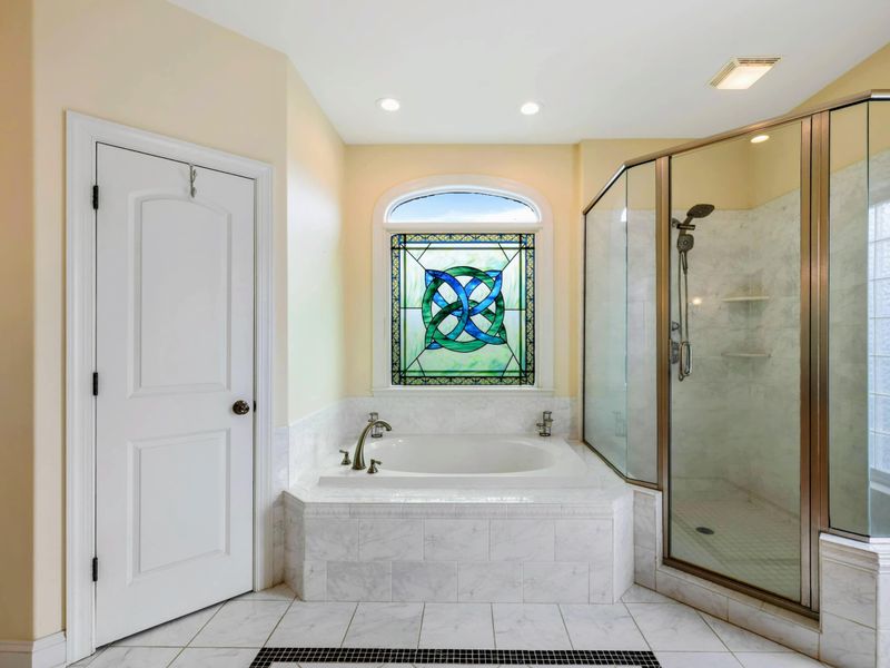 Primary Ensuite - A soaking tub beneath a stained glass window, with a glass-enclosed walk-in shower and marble tile throughout.