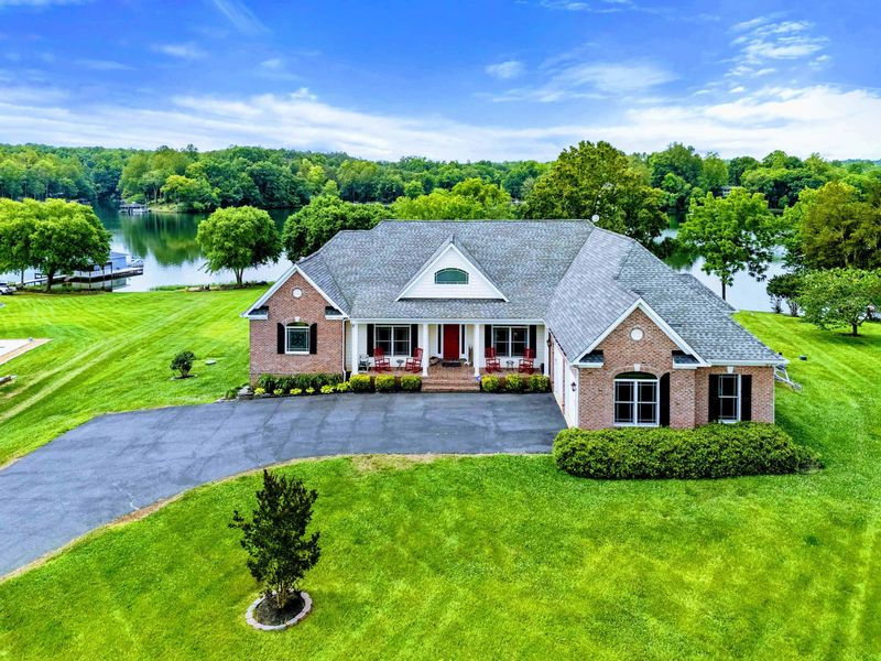 Aerial View - A generous green lot with circular driveway leads to the brick home, with the lake stretching out just behind.