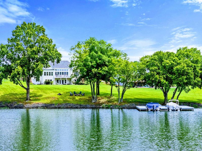 From the Water - Mature trees line 220 feet of shoreline, with the home and private dock set back on a lush green lawn.