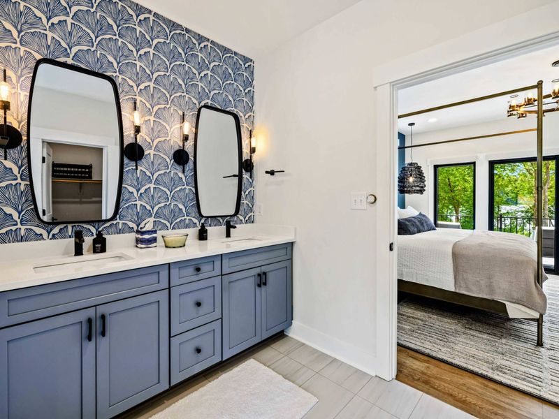 Primary Bathroom - A double vanity with blue cabinets and ginkgo leaf wallpaper connects to the canopy bedroom through a wide doorway.