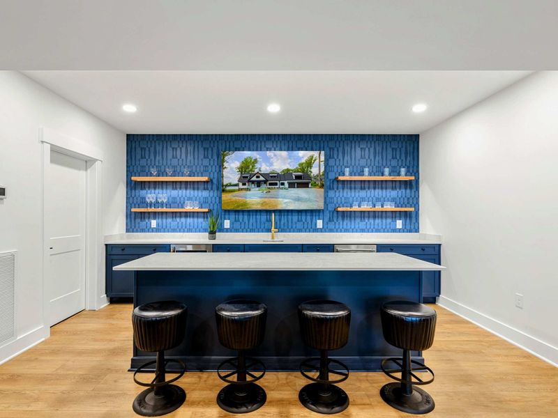 Wet Bar - Four swivel stools line the island beneath a gold faucet and floating shelves stocked with glassware.