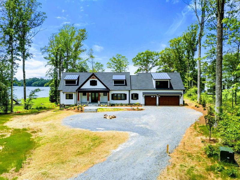 Aerial View - The home and attached garage sit on a wooded lakefront lot with the boathouse and sandy beach visible beyond.