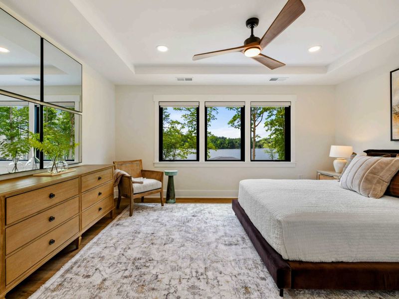 Bedroom 6 - A king bed with earth-tone bedding and plaid accent pillows sits beneath a ceiling fan with three windows framing lake views.