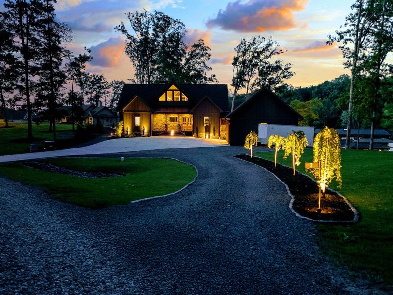 Driveway Approach - The winding gravel drive leads through uplighted weeping trees to the illuminated home, creating a dramatic arrival at sunset.