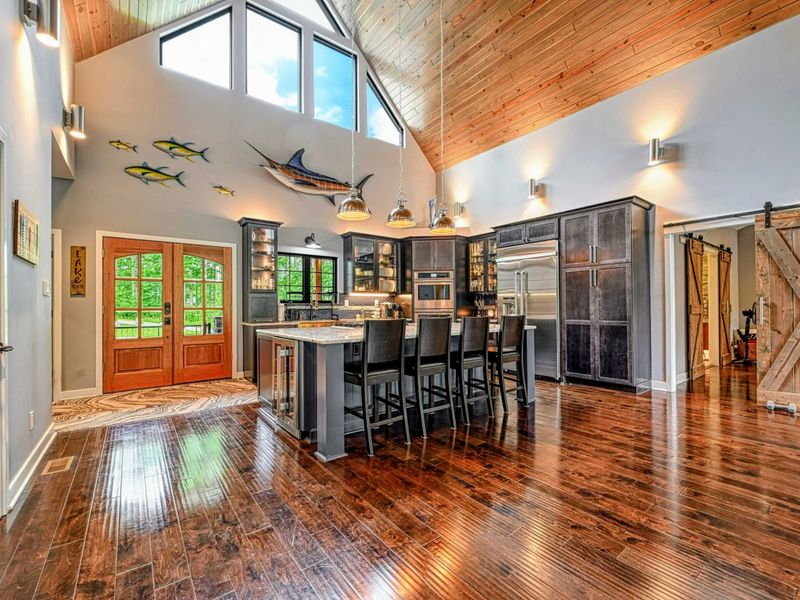 Open Floor Plan - The kitchen island, bar stools, and great room flow together beneath the pine cathedral ceiling, with the front entry and barn doors in view.