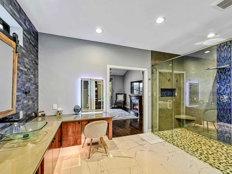 Primary Bathroom - A spa-inspired en suite with glass vessel sinks on a cherry wood vanity, stacked stone accent wall, pebble-floor shower with blue mosaic tile, and a vanity desk.