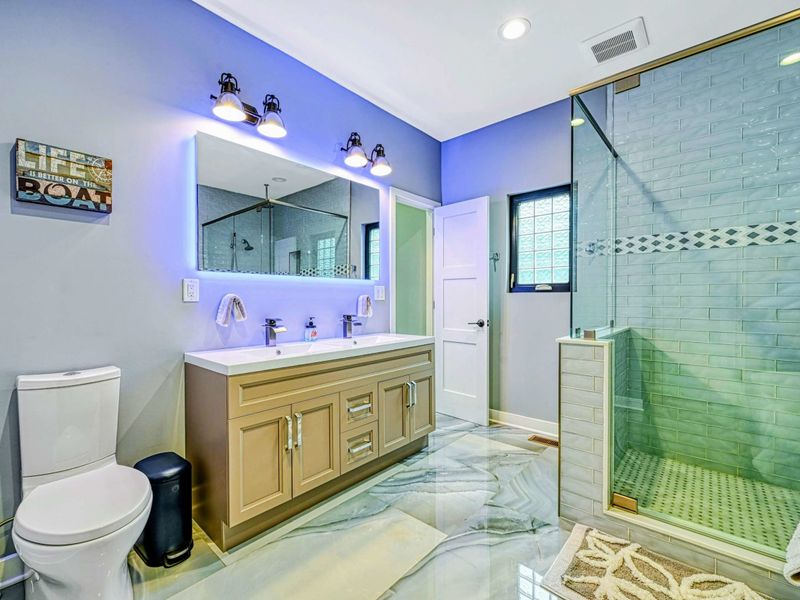 En Suite Bathroom - Another view of the spa bathroom showing the double vanity, glass shower, and marble-look floors under soft purple-blue ambient lighting.