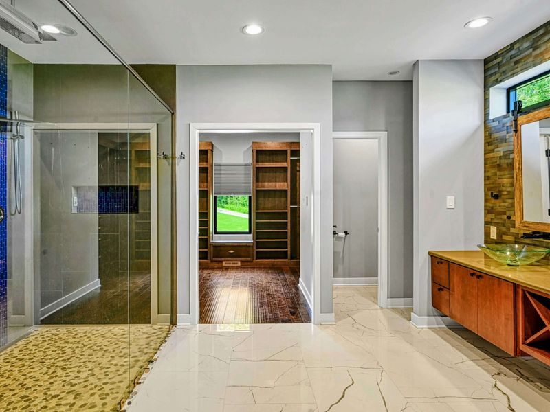 Primary Bathroom - The full en suite showing the oversized glass-enclosed shower, pebble flooring, blue mosaic accents, walk-in closet entry, and cherry vanity.