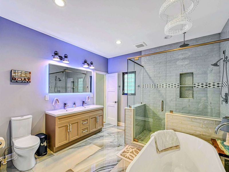 En Suite Bathroom - A luxurious spa bath with LED-backlit mirror, double vanity, glass-enclosed walk-in shower with decorative tile, freestanding soaking tub, and crystal chandelier.