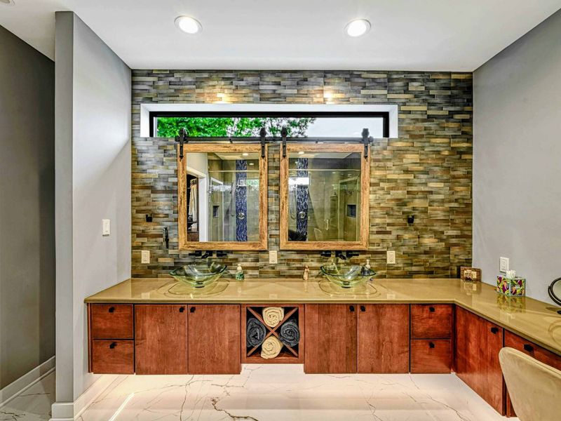 Primary Double Vanity - Dual glass vessel sinks on a cherry vanity with barn door mirrors, stacked stone backsplash, and a view through to the bedroom fireplace.