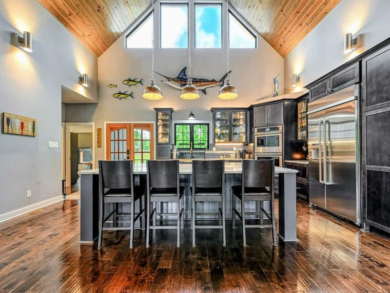 Kitchen - Six black leather stools line the oversized granite island beneath industrial pendant lights, with the soaring A-frame ceiling and marlin wall art above.