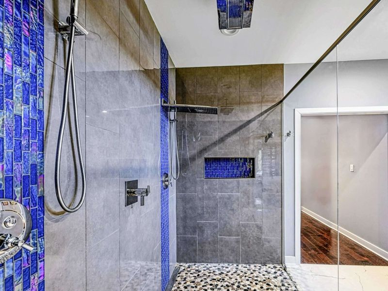 Primary Shower Detail - A close-up of the dual shower heads, blue mosaic tile accents, and pebble floor inside the oversized walk-in shower.
