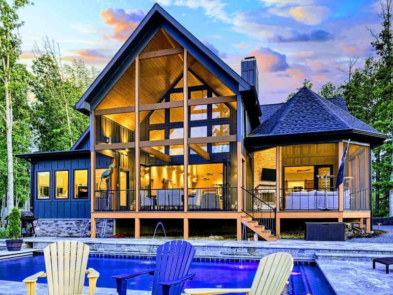 Twilight Rear View - The full lakeside exterior at golden hour, with LED-lit pool, Adirondack chairs, and the screened porch glowing beyond.