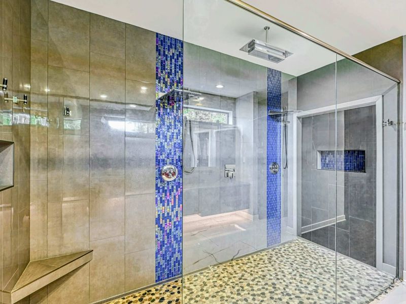 Primary Shower - A massive glass-enclosed shower with rainfall and handheld heads, pebble stone floor, blue mosaic tile columns, and a corner bench.