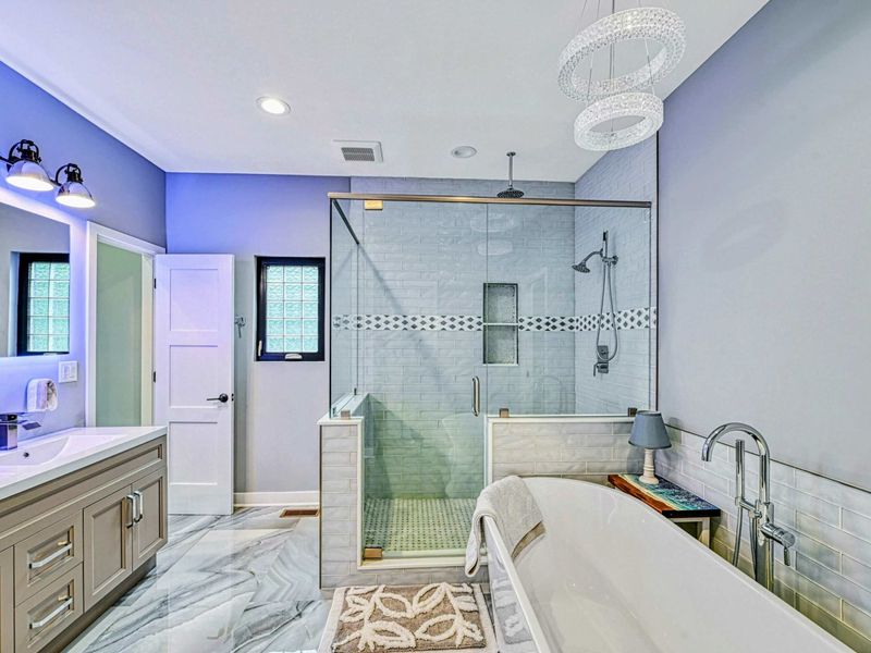 Soaking Tub - A freestanding soaking tub beneath a crystal chandelier, beside the glass-enclosed shower with decorative tile accents and a marble-look floor.