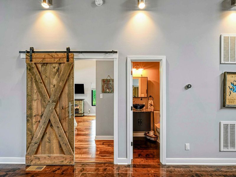 Hallway - A rustic X-pattern barn door on a sliding track leads to a bedroom, with a powder room entry and Lake Anna wall art alongside.
