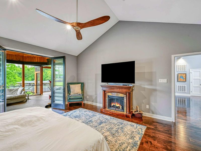 Primary Suite - Vaulted ceilings, a stone fireplace, Smart TV, accent chair, and French doors to the screened porch with lake views beyond.