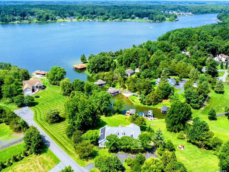 Aerial - Neighborhood perspective showing the home, green lawns, mature trees, and proximity to the lake.