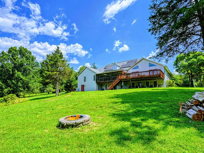 Exterior - Rear of the home with a stone fire pit in the lawn, wraparound deck, and solar panels on the roof.