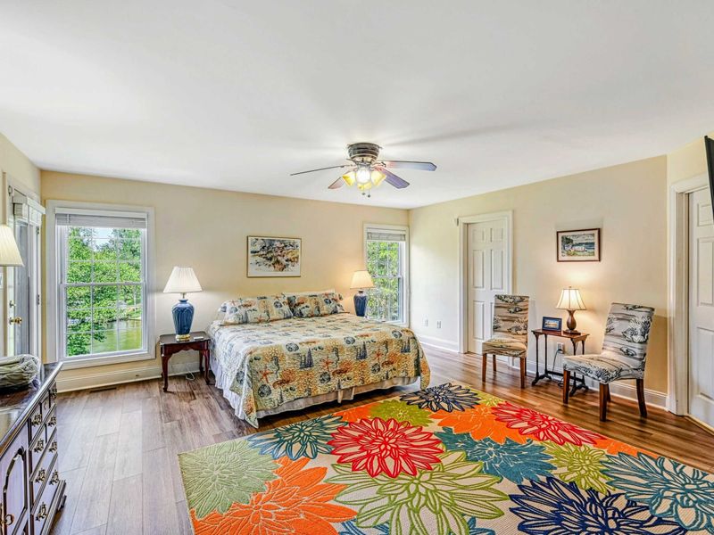Primary Suite - King bed with floral bedding, colorful area rug, ceiling fan, TV, and lake views through the windows.