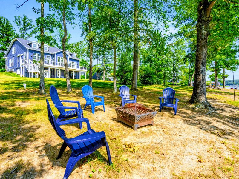 Fire Pit Area - Blue Adirondack chairs ring the fire pit on the shaded lawn, with the home and lake framing the scene.